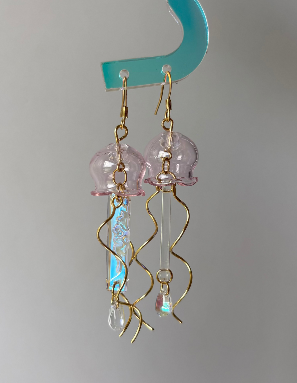 A pair of handmade jellyfish dangle earrings held by a turquoise plastic hanger. The earrings feature light pink translucent jellyfish charms, gold fishhook wires, and iridescent blue-green wavy tails with small clear crystal drops.
