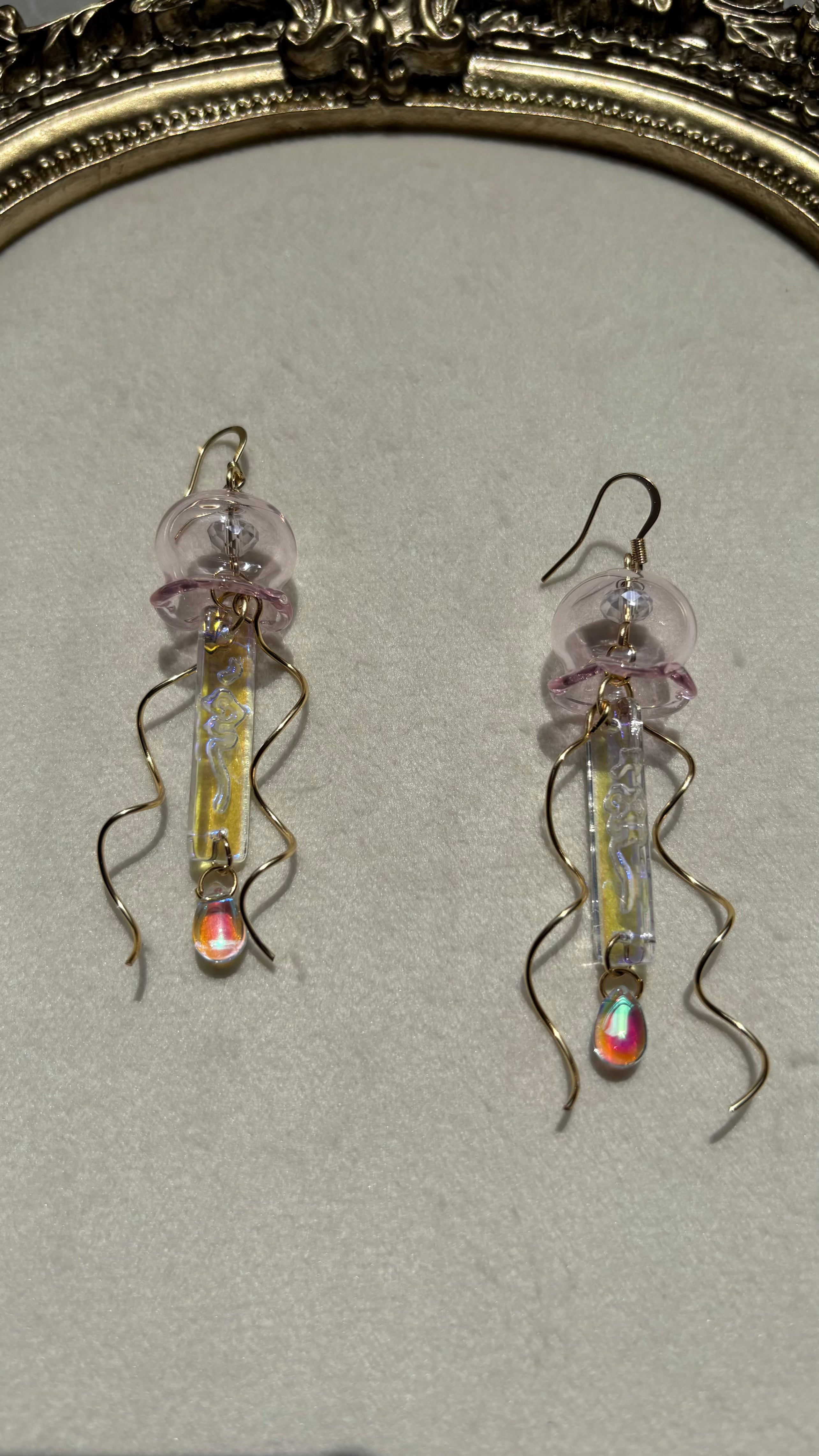 A pair of handmade jellyfish-inspired dangle earrings displayed on a neutral textured surface with an ornate gold frame in the background. The earrings feature a translucent pink jellyfish head, a vertical iridescent yellow tube, and wavy gold tentacles ending in small iridescent teardrop charms.